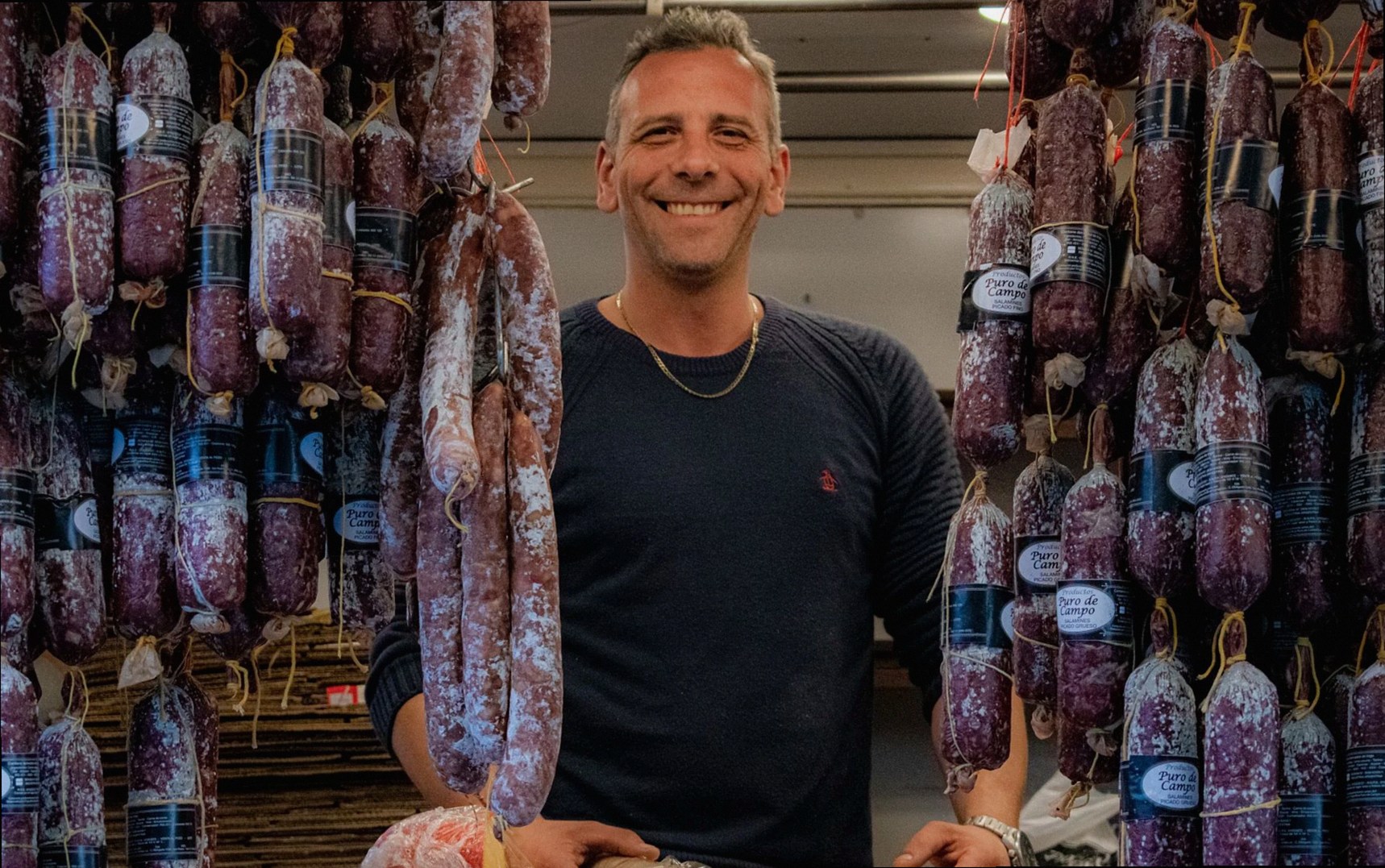 Vendeur souriant posant avec des saucissons suspendus dans un marché local en Argentine, mettant en valeur la culture cu